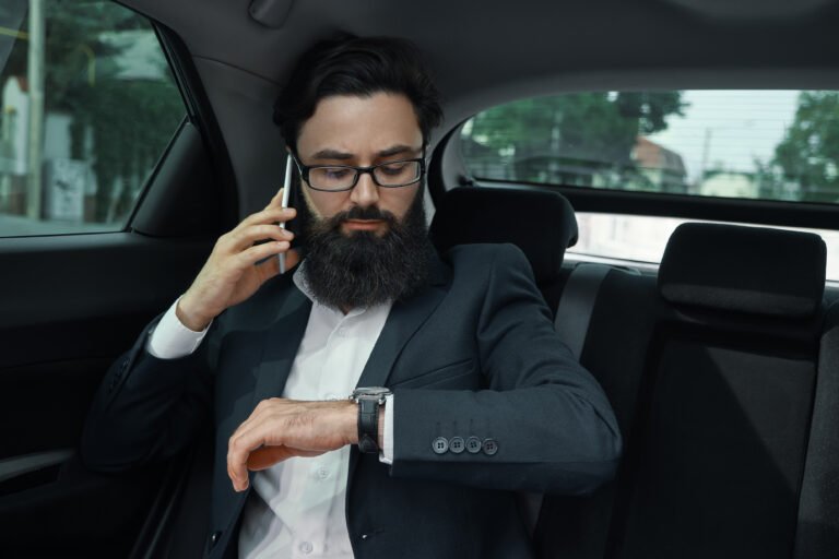 A businessman while traveling by car in the back seat. A handsome young man in full suit looking, talking on his smartphone and looking at his watch while sitting in the car.